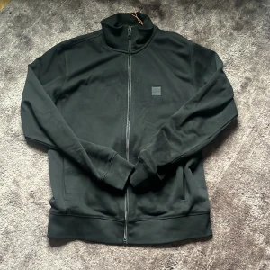 Svart ziptröja från BOSS med orange detalj - Svart långärmad ziptröja från BOSS med hög krage och diskret logotyp på bröstet. Snygg orange detalj på insidan av kragen. Tröjan är i mjukt bomullsmaterial och har en clean look med dragkedja framtill.