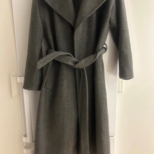 Grå lång kappa med bälte - Säljer en stilren grå kappa med bred krage och knytbälte i midjan. Modellen är lång och har raka ärmar, perfekt för dig som gillar minimalistisk och clean look. Materialet känns mjukt och har en ullig finish. Snygg att slänga över vilken outfit som helst.