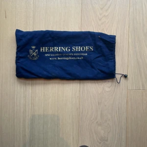 Svarta skinnskor från Herring Shoes - Klassiska svarta skinnskor från Herring Shoes med snygga broguedetaljer och snörning. Skorna har rund tå och låg klack, perfekt för en dressad look. Levereras med originalpåse. Stilrena och tidlösa, passar till kostym eller uppklädda tillfällen.