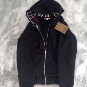 Svart Burberry hoodie med rutig huva - Svart hoodie från Burberry med klassisk rutig insida i huvan i svart, vit och röd. Tröjan har dragkedja framtill, snörning vid huvan och två fickor. Perfekt för dig som gillar stilrena detaljer och ikoniskt mönster. Tvekan inte på att fråga!✅