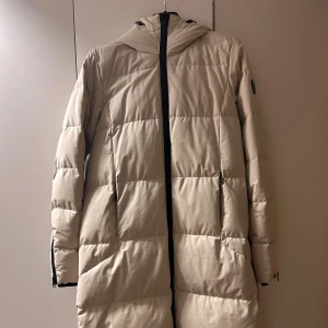 Beige lång dunjacka från Tenson - Säljer en beige lång dunjacka från Tenson med huva och svarta dragkedjor. Jackan har quiltad design och är perfekt för kalla dagar. Den har praktiska fickor med dragkedja och en snygg logga i nacken. Passformen är normal och ärmarna är långärmade. Väldigt få använd.