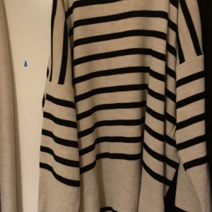 Beige stickade mjukisbyxor o tröja  -  Ett ljust beige eller gråbeige plagg, troligen ett par stickade byxor eller en lång kofta. Tyget ser mjukt och lite tjockare ut . En tröja i liknande ljus färg men med mörkblå eller svarta horisontella ränder. Den har en polo- eller polokrage och vida ärmar, vilket ger ett mysigt och lite marint intryck.