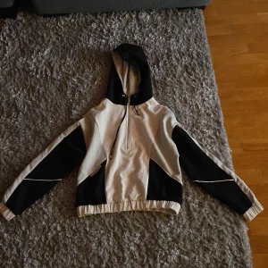 Svartvit vindjacka med luva  - Säljer en svartvit vindjacka med coola paneler och half zip framtill. Jackan har luva med snörning och elastisk nederkant för skön passform. Materialet känns som syntet och polyester, perfekt för blåsiga dagar. Långärmad och loose fit för chill stil.