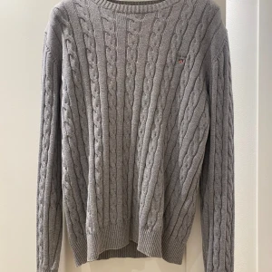 Grå kabelstickad tröja från Gant - Snygg grå kabelstickad tröja från Gant i premium bomull. Tröjan har rund hals, ribbade muddar och ett broderat Gant-emblem på bröstet. Perfekt för dig som gillar klassisk stil med en modern touch.