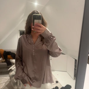 Silvrig satinblus från Stockh LM - Säljer en silvrig satinblus från Stockh LM med v-ringning och snygga pärlemorknappar framtill. Blusen har långa ärmar med breda manschetter och en lös, flowy passform. Perfekt för dig som gillar en stilren och trendig look.