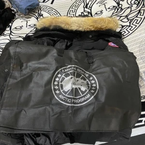 Svart dunjacka Canada Goose med päls - Svart dunjacka från Canada Goose med ikonisk röd-vit-blå patch på ärmen och fluffig beige päls runt huvan. Jackan har dragkedja, knappar och quiltad design. Perfekt för kalla vinterdagar och har en clean, klassisk look.