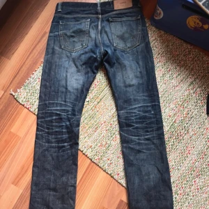Edwin selvedge jeans - Mörkblå edwin jeans i selvedge denim med klassisk rak passform. Snygga slitningar och en liten reva på ena benet ger en cool, använd look. Femficksmodell med knappar i gylfen och kontrastsömmar. Perfekta för dig som gillar autentisk denimstil.