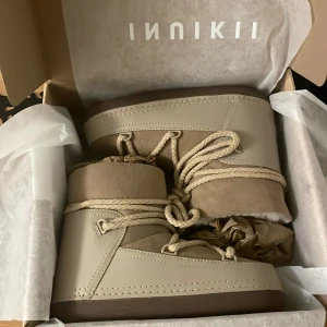 Beige vinter skor från Inuikii - Säljer ett par snygga beige vinter skor från Inuikii med snören uppepå och mjuk insida i vit päls. Skorna har en rund tå, platt sula och är tillverkade i skinn och mocka. Perfekta för kalla dagar och ger en cool vinterlook. ENDAST PROVADE INOMHUS!!