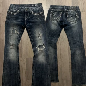 Miss Me blå bootcut jeans med nitar - Snygga blå jeans från Miss Me med bootcut passform. Jeansen har slitna detaljer, hål och fransar på benen samt nitar och glittriga stenar på fickor och kanter. Tuffa kontrastsömmar och dekorativa knappar ger extra edge. Perfekta för dig som gillar statement-jeans.