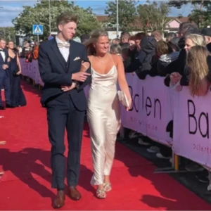 Champagnefärgad långklänning i satin - Säljer en elegant långklänning i champagnefärgad satin med tunna axelband och öppen rygg. Klänningen har en smickrande v-ringning och följsam passform som ger en lyxig känsla. Perfekt för bal eller student! 