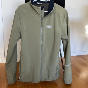 Grön vindjacka från Helly Hansen - Snygg grön softshell från Helly Hansen med HH-logga på bröstet. Jackan har hel dragkedja framtill, hög krage och är fodrad med mjuk fleece på insidan. Långärmad modell med diskreta reflexdetaljer på ärmarna. Perfekt för blåsiga dagar och outdoor. Jackan är använd vid enstaka tillfällen, så i nyskick. 