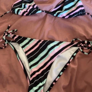 Randig bikini i pastellfärger - Säljer en snygg bikini med svarta, mintgröna, rosa och vita ränder. Överdelen är trekantsmodell med knyt i nacken och pärldetaljer framtill. Underdelen har knyt i sidorna. Perfekt för stranden eller poolen i sommar!