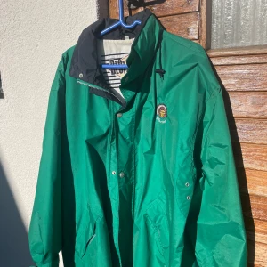 Grön vindjacka från Blount Proud - Snygg grön vindjacka från Blount Proud med svart krage och coolt broderat emblem på bröstet. Jackan har dragkedja, knappar och praktiska fickor samt snörning vid kragen. Perfekt för dig som gillar streetstyle och vill sticka ut.