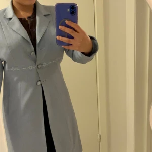 Ljusblå långklänning med broderi - Elegant ljusblå långkappa/rock med klassisk krage, långa ärmar och dekorativa broderier vid midjan och ärmsluten. Klänningen har tre klädda knappar framtill och är sydd i ett mjukt, vävt material som ger en stilren look.