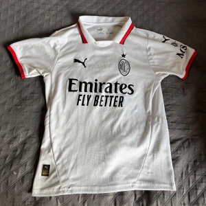 Vit AC Milan fotbollströja Puma M - AC Milan bortatröja från Puma i vitt med svarta och röda detaljer. Broderat klubbmärke och stjärna på bröstet, svart Puma-logga och sponsortryck. Tillverkad i lätt, ventilerande material med korta ärmar och klassisk krage.