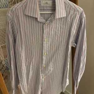 Randig skjorta från Carolina Herrera - Snygg skjorta från Carolina Herrera med tunna röda ränder på vit botten. Klassisk krage, långa ärmar och knappar med logotyp. Tillverkad i Portugal av mjuk bomull. Perfekt för dig som gillar stilrena och tidlösa plagg med en twist. Obs, fläck på andra bilden som går att få bort med fläckborttagningsmedel eller galltvål.