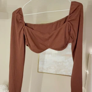 Brun långärmad korsett-topp - Säljer en snygg brun korsett-topp med lång ärm och fyrkantig urringning. Toppen har en ribbad struktur och markerad byst för en trendig look. Perfekt att styla med höga jeans eller kjol 