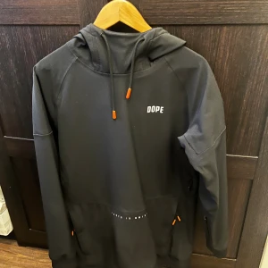 Jacka från Dope - Svart jacka från Dope med huva och orange detaljer på snören och fickor. Långärmad med tryckt logga på bröstet och patch på ärmen. Praktisk ficka med dragkedja framtill. Har även en ficka på armen för liftkort. Priset är ej fast