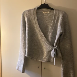 Grå omlottkofta från 157 XS - Mysig grå omlottkofta från 157 i storlek XS. Koftan har lång ärm, ribbade muddar och knyt i sidan som ger en snygg och avslappnad look. Perfekt för lager-på-lager och chill dagar. Materialet är stickat och känns mjukt mot huden.