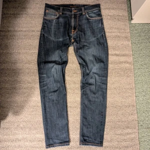 Nudie Jeans - Ett par väldigt fina Nudie Jeans i modellen Thin Finn storlek 32/32. Passformen är slim. De har den klassiska brodyren i vitt på bakfickorna och orangea sömmar. Enda tecken på användning är mindre slitningar i sömmarna till fickorna och i bensluten, samt mindre färgavvikelser efter tvätt. Hör gärna av er om det är något ni undrar över! Färgen är inte precis som på bilden med tanke på olika ljus .