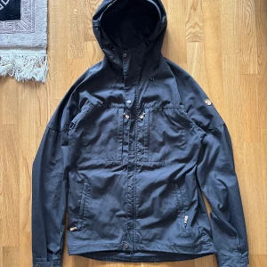 Fjällräven svart skogsö jacka, storlek M - Svart jacka med huva, dragkedja och knappar framtill. Jackan har flera fickor med dragkedja och knappar. Justerbar huva med dragsko. Logotyp på vänster ärm.