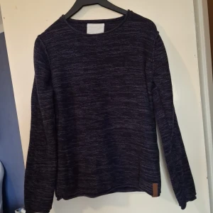 Mörkblå stickad tröja med rund hals - Mysig mörkblå stickad tröja med diskret melerat mönster och rund halsringning. Tröjan har långa ärmar och en liten brun detalj nertill. Perfekt för lager-på-lager under kyliga dagar.