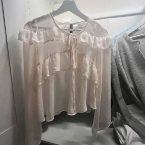 blus med volanger från H&M - Supersöt ljusrosa blus från H&M med volanger och knyt framtill. Blusen har offshoulder-design och är tillverkad i ett tunt, flowy material som ger en romantisk vibe. Perfekt för dig som gillar feminina detaljer och en luftig känsla.
