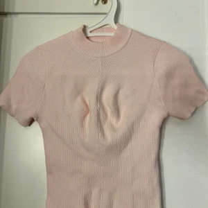 Ljusrosa ribbad topp - Supersöt ljusrosa ribbad topp från Pomelo med kort ärm och hög krage. Klänningen har en figurnära passform och är tillverkad i ett stretchigt, stickat material som ger en snygg siluett. Perfekt för dig som gillar en enkel men trendig look.