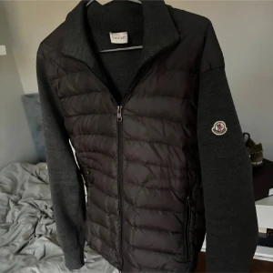 Moncler cardigan  - Snygg grå cardigan jacka från Moncler med quiltad framsida och stickade ärmar och rygg. Jackan har dragkedja framtill, hög krage och Moncler-logga på ärmen. Perfekt för dig som vill ha en stilren och exklusiv look. Äkthets verifierad av Vestiaire Collective och denna färg säljs inte mer. Lite skavanker på slutet av ärmarna.