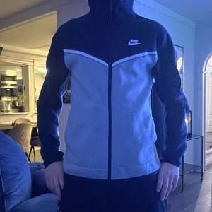 Nike svart och grå vindjacka - Nike vindjacka med huva i svart och ljusgrått. Jackan har hel dragkedja framtill, två sidofickor och en vit detalj längs bröstet. Perfekt för vardagsstil och sportiga outfits.