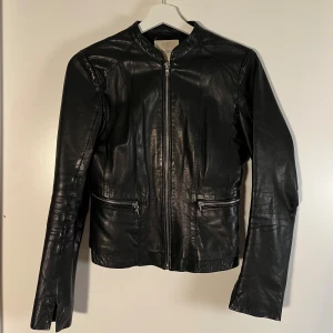 Svart skinnjacka från Mango XS - Snygg svart skinnjacka från Mango i äkta läder. Jackan har en stilren design med raka linjer, silverfärgad dragkedja framtill och två fickor med dragkedja. Långärmad och figurnära passform som ger en cool vibe till din outfit. Det finns en liten defekt (2 cm) på baksidan av höger ärm.