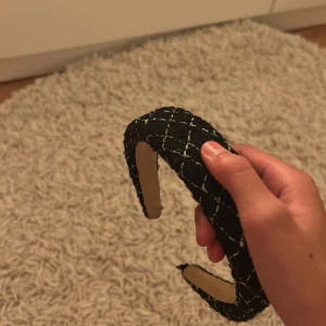 Svart rutigt diadem i tweed - Snyggt svart diadem med vit rutigt mönster, tillverkat i tweed-liknande tyg. Diademet är brett och har en mjuk, vadderad känsla som ger en cool och trendig look. Perfekt för att lyfta din outfit med en chic touch.