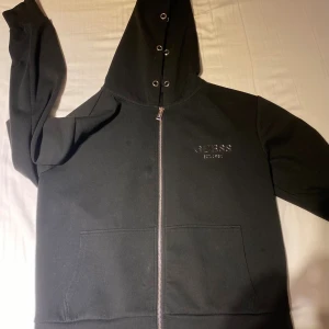 Svart hoodie med dragkedja från Guess - Svart hoodie från Guess med dragkedja framtill, huva med metallöljetter och broderad logga på bröstet. Tröjan har känguruficka och är tillverkad i mjukt material som känns skönt mot huden. Perfekt för en chill och stilren look.