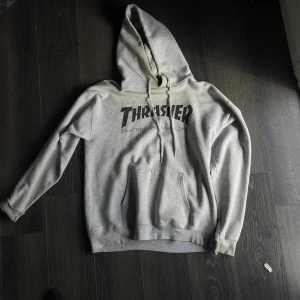 Grå Thrasher hoodie med tryck - Säljer en grå hoodie från Thrasher med klassiskt svart logotryck på bröstet. Tröjan har huva med snörning och en stor magficka. Perfekt för dig som gillar streetwear och skatekänsla.