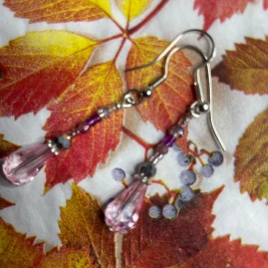  Hangjorda, långa örhängen med rosa droppar - Säljer ett par snygga handgjorda örhängen med silver s 925 krokar och hängande pärlor i  rosa och lila toner. De har en droppformad rosa pärla längst ner och små glittriga detaljer längs hela hänget. Perfekta för dig som gillar färg och vill sticka ut lite extra. De stora dropparna är facettslipatglas de små (runda) halvädelstenar.