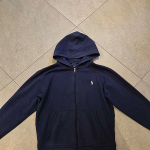 Marinblå hoodie från Polo Ralph Lauren - Säljer en marinblå hoodie från Polo Ralph Lauren med dragkedja och klassisk broderad logga på bröstet. Tröjan har luva, fickor framtill och är tillverkad i mjuk bomull. Perfekt för en chill och stilren look.