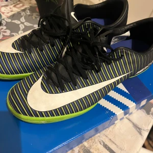 Nike Mercurial randiga fotbollsskor - Säljer ett par Nike Mercurial fotbollsskor med svart bas, tunna blå och neongröna ränder samt stora vita swoosh-loggor. Skorna har snörning och platt sula, perfekt för inomhusspel. Materialet är syntet och insidan är fodrad i blått. Riktigt snygga och sportiga skor för dig som älskar fotboll.