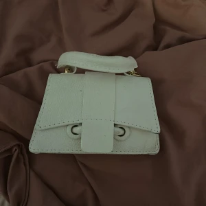 Vit minihandväska i skinn - Superfin liten handväska i vitt skinn med stilren design och guldfärgade detaljer. Väskan har ett kort handtag upptill och stängs med magnetknappar. Perfekt för att bära det viktigaste och ger en clean look till din outfit.