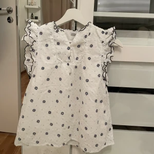 Vit blus med små blå blommor & puffärmar – Lindex - Söt vit blus från Lindex med små blå blommor och puffärmar. Lätt och mjuk bomullskvalitet som känns skön mot huden. Knappslå framtill och som passar både till vardag och fest. Storlek 158/164. Mycket fint skick – knappt använd och ser nästan ny ut. (Andvänd 1 gång)