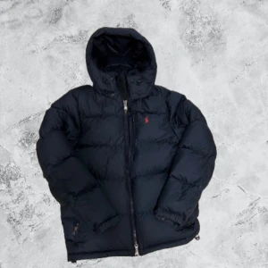 Mörkblå från Polo Ralph Lauren - Snygg mörkblå dunjacka från Polo Ralph Lauren med huva och dragkedja. Jackan har en röd broderad logga på bröstet, praktiska fickor med dragkedja och quiltad design. Perfekt för kalla dagar och har en clean, sportig look. Använd, men skicket är nästan lika bra som ny. Priset kan sänkas lite vid snabb affär. Skriv vid   frågor!