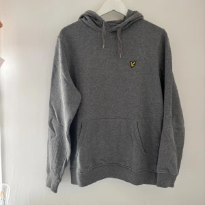 Grå hoodie från Lyle & Scott - Säljer en grå hoodie från Lyle & Scott med klassisk gul fågel-logga på bröstet. Tröjan har huva med snörning, magficka och ribbade muddar. Perfekt för chill dagar och enkel att matcha med jeans eller joggers.
