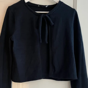 Svart kofta med knyt framtill - Snygg svart kofta med lång ärm och knytband framtill vid halsen. Enkel och stilren design som funkar till många olika looks. Mjuk och skön kvalitet, perfekt att slänga på sig när det är lite kyligt ute.