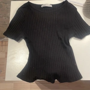 Svart ribbad omlottopp FB Sister XS - Svart ribbad omlottopp från Newyorker i storlek XS. Toppen har korta ärmar, rund halsringning med en halvt öppen rygg och knytband i midjan för en snygg passform. Perfekt att styla med jeans eller kjol för en trendig look.