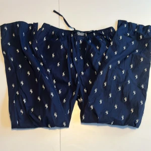 Pyjamasbyxor Polo Ralph Lauren - Mörkblå pyjamasbyxor från Polo Ralph Lauren med vitt broderat logomönster över hela byxan. De har resår och snörning i midjan för skön passform. Tillverkade i 100% bomull. Helt oanvända pågrund av att de är för stora för mig