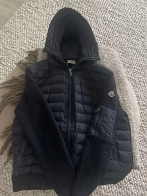 Marinblå Monclerjacka med huva - Riktigt snygg jacka från Moncler med huva och dragkedja framtill. Jackan har quiltade paneler och stickade partier på ärmar och rygg, liten ficka på ena ärmen samt sidorna.                                     Storlek XL, men passar M-XL