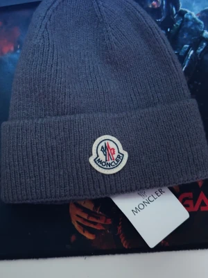 Grå stickad Moncler mössa - Snygg grå stickad mössa från Moncler med klassisk logotyp framtill. Mössan har en bred uppvikt kant och är tillverkad i mjukt material som håller dig varm. Perfekt accessoar för kyliga dagar och enkel att matcha med din outfit.