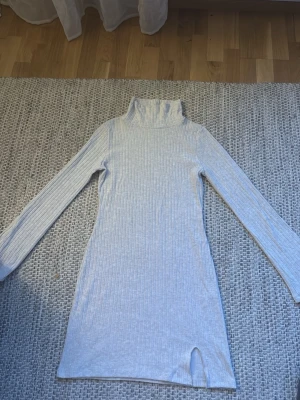 Ljusgrå ribbad poloklänning - Säljer en ljusgrå ribbad klänning med polokrage och långärmade ärmar. Klänningen är kort och har en liten slits framtill. Materialet känns mjukt och stretchigt. 