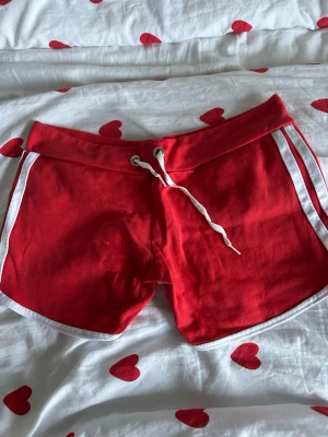 Röda sportiga shorts med vita ränder - Säljer ett par röda shorts med vita ränder längs sidorna och vit snörning i midjan. De har en sportig look med rundade kanter och vit kant runt benen. Materialet känns mjukt och är troligtvis bomull eller polyester. Perfekta för sommarens aktiviteter!