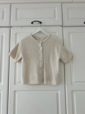 Beige stickad kofta från Gina Tricot💖 - Kortärmad beige stickad kofta från Gina Tricot med rund hals och ribbad struktur. Koftan har knappar framtill i pärlemor och är tillverkad i en mjuk ullblandning. Perfekt för lager-på-lager och chill stil.