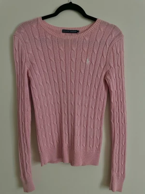 Rosa kabelstickad tröja Ralph Lauren - Supersöt kabelstickad tröja från Ralph Lauren i ljusrosa. Klassisk rund halsringning, långärmad och med diskret logga broderad på bröstet. Perfekt till jeans eller kjol för en clean och preppy look😍 I nytt skick, enbart testad en gång. 